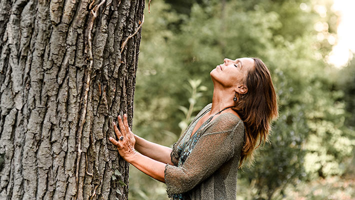 Frau steht mit geschlossenen Augen an einem Baum und berührt ihn während einer Selbsthypnose